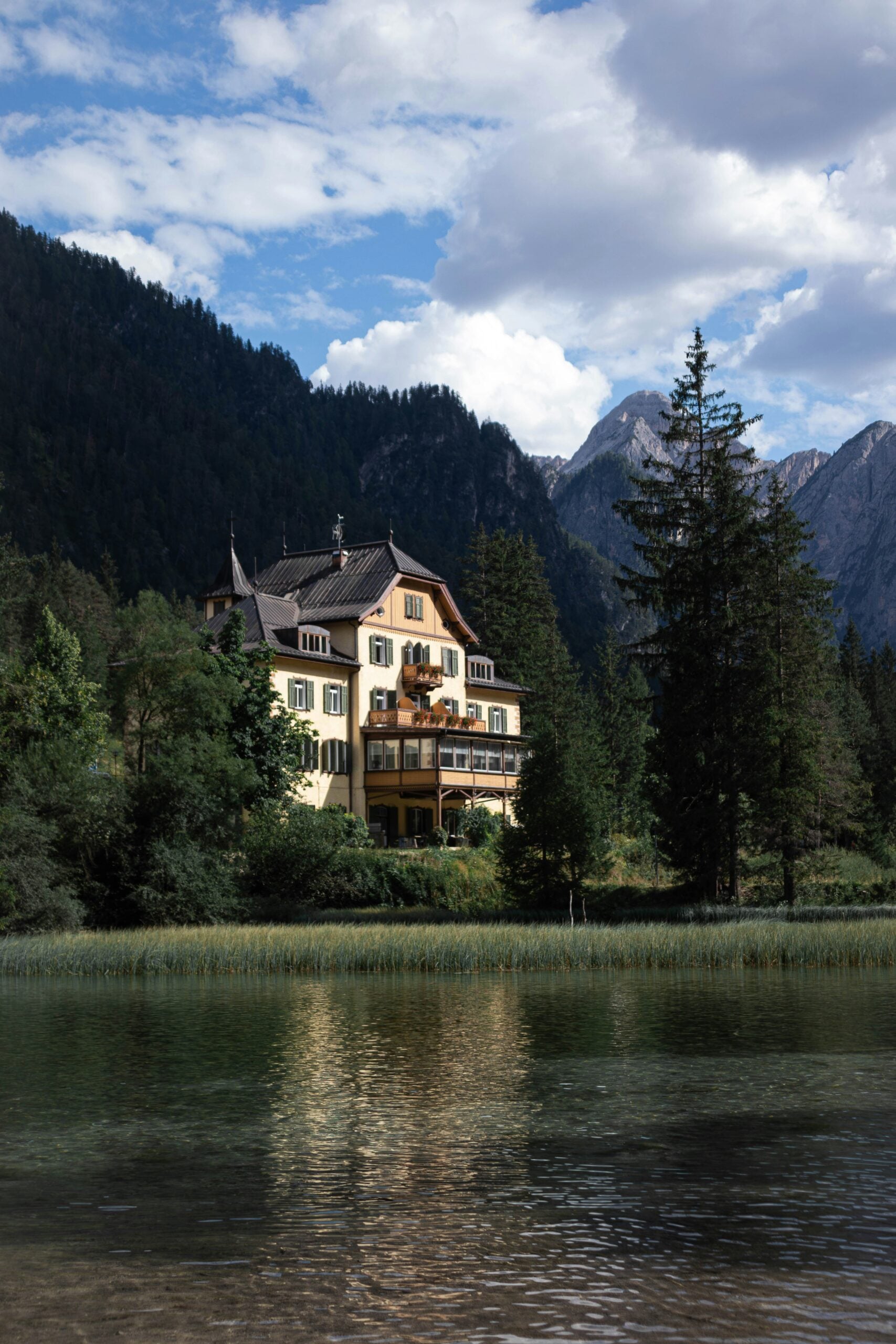 A picturesque scene of a charming house nestled by the edge of a tranquil lake in Bolzano, Italy. The house, with its traditional Alpine architecture, features wooden beams, flower-adorned balconies, and a sloping roof, perfectly complementing the serene natural surroundings. The calm lake reflects the house and the surrounding lush greenery, creating a mirror-like surface that adds to the peaceful ambiance. In the background, the rolling hills and distant peaks of the Dolomites rise gracefully, completing this idyllic setting. This image beautifully captures the harmonious blend of architecture and nature that Bolzano, Italy, is renowned for.