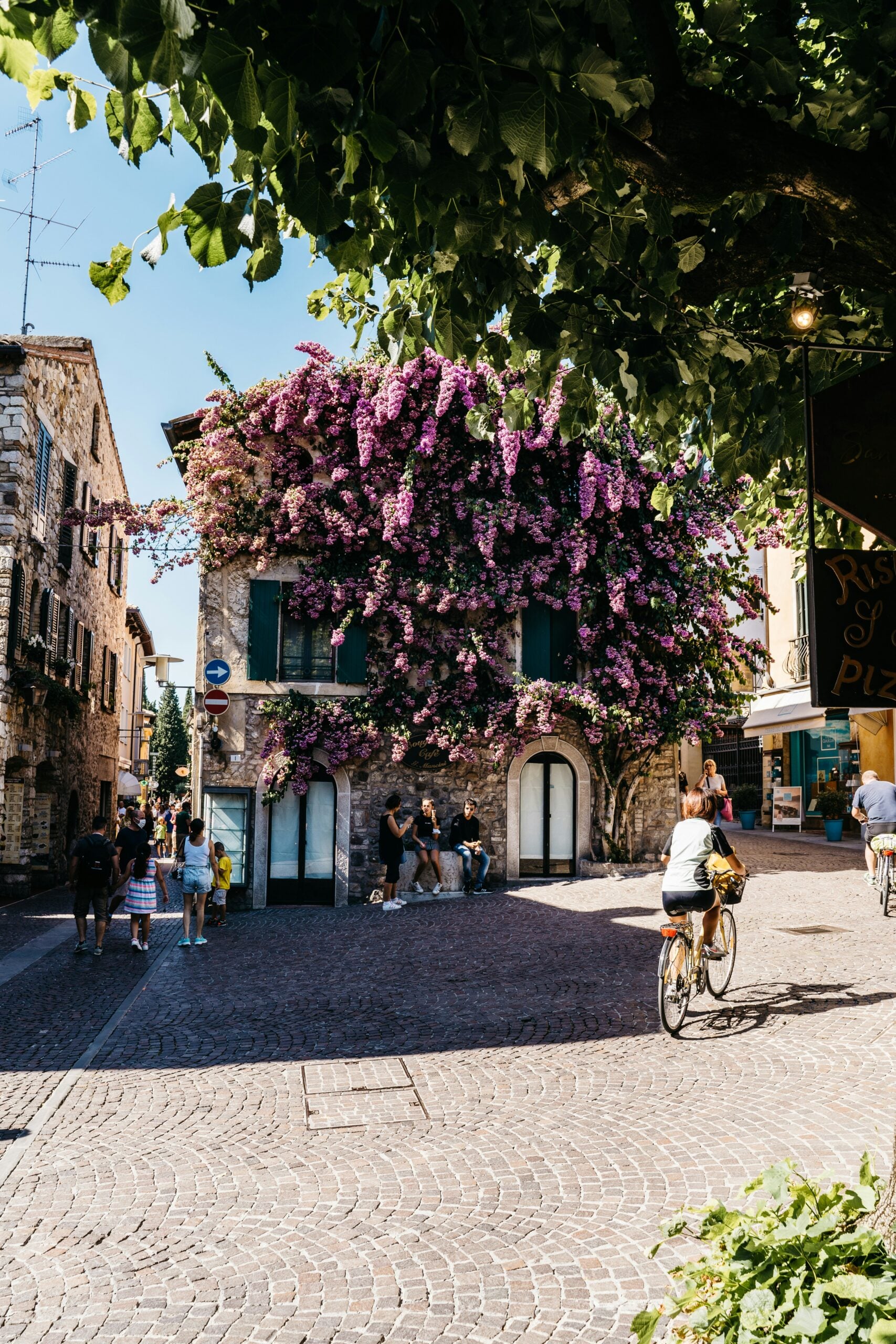 Photograph of a charming street scene in Sirmione, located in the Province of Brescia, Italy. This image captures the quintessential cobblestone streets lined with vibrant flowers and quaint local shops, inviting visitors to stroll and explore. The historic buildings and relaxed atmosphere provide a picturesque setting, embodying the enchanting character of this Italian lakeside town.