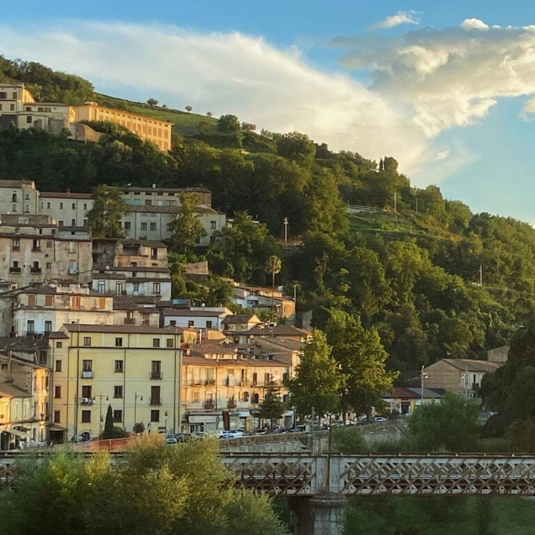 A hillside town in Calabria, with pastel-colored buildings and rooftops nestled among lush green trees under a partly cloudy sky. In Cosenza, a metal bridge crosses in the foreground, all bathed in warm, golden sunlight.
