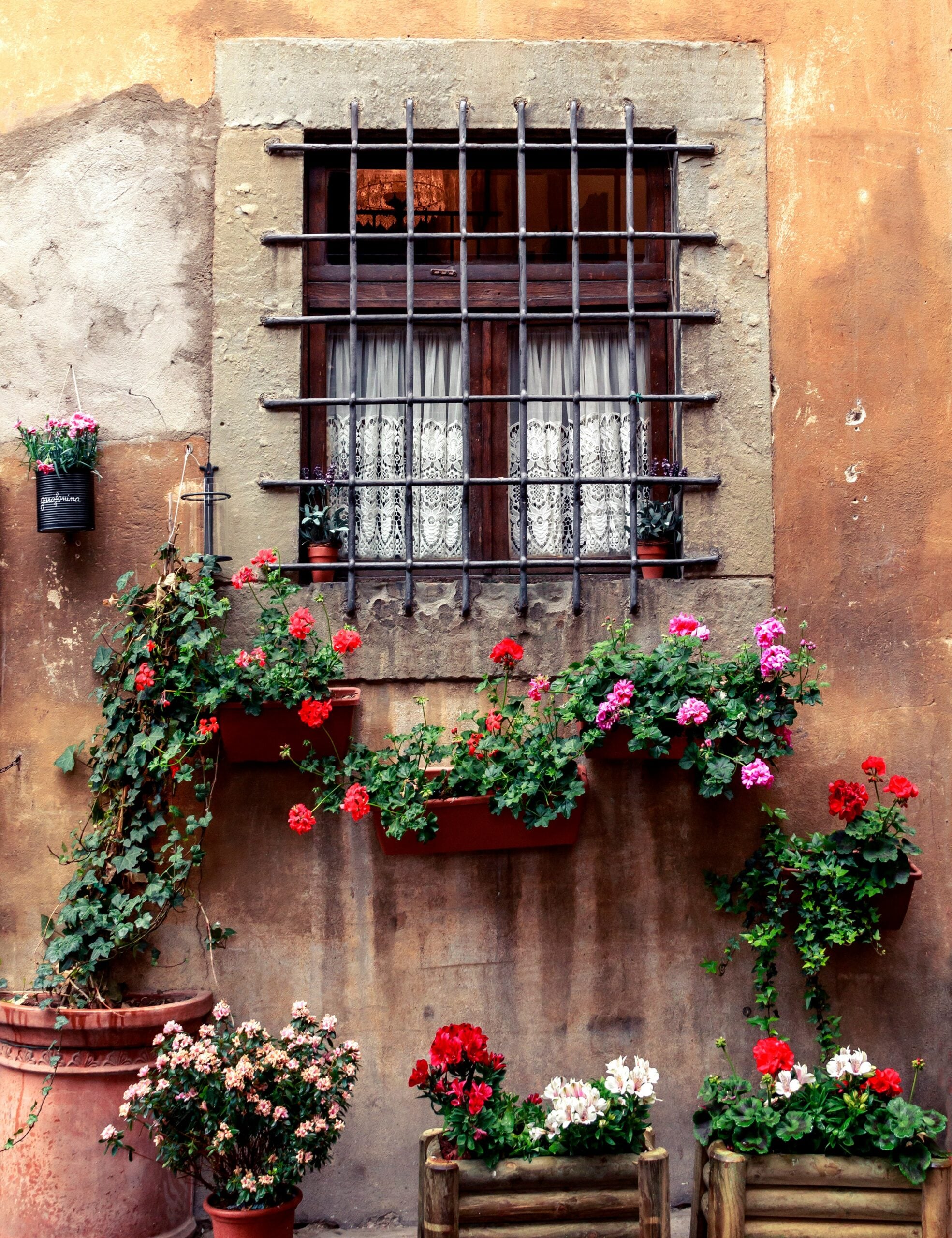The image shows a charming window set into an old, weathered wall of a residence, likely in a historic area. The wall is constructed of aged stone or stucco, showing the texture and character that comes with time. The window is framed by wooden shutters, painted in a faded color that complements the rustic look of the wall. A vibrant display of flowers in a window box or planter adds a splash of color and life to the scene, with blooming plants cascading over the edges. The overall effect is one of quaint beauty, capturing the timeless charm of a traditional, well-loved home in a historic setting.