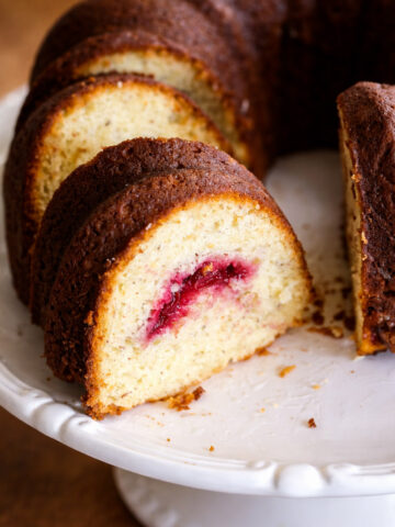 A sliced Lemon Cranberry Cake sits on a white cake stand, revealing a layer of red fruit filling inside the moist yellow cake. Crumbs are scattered on the stand next to the cake.