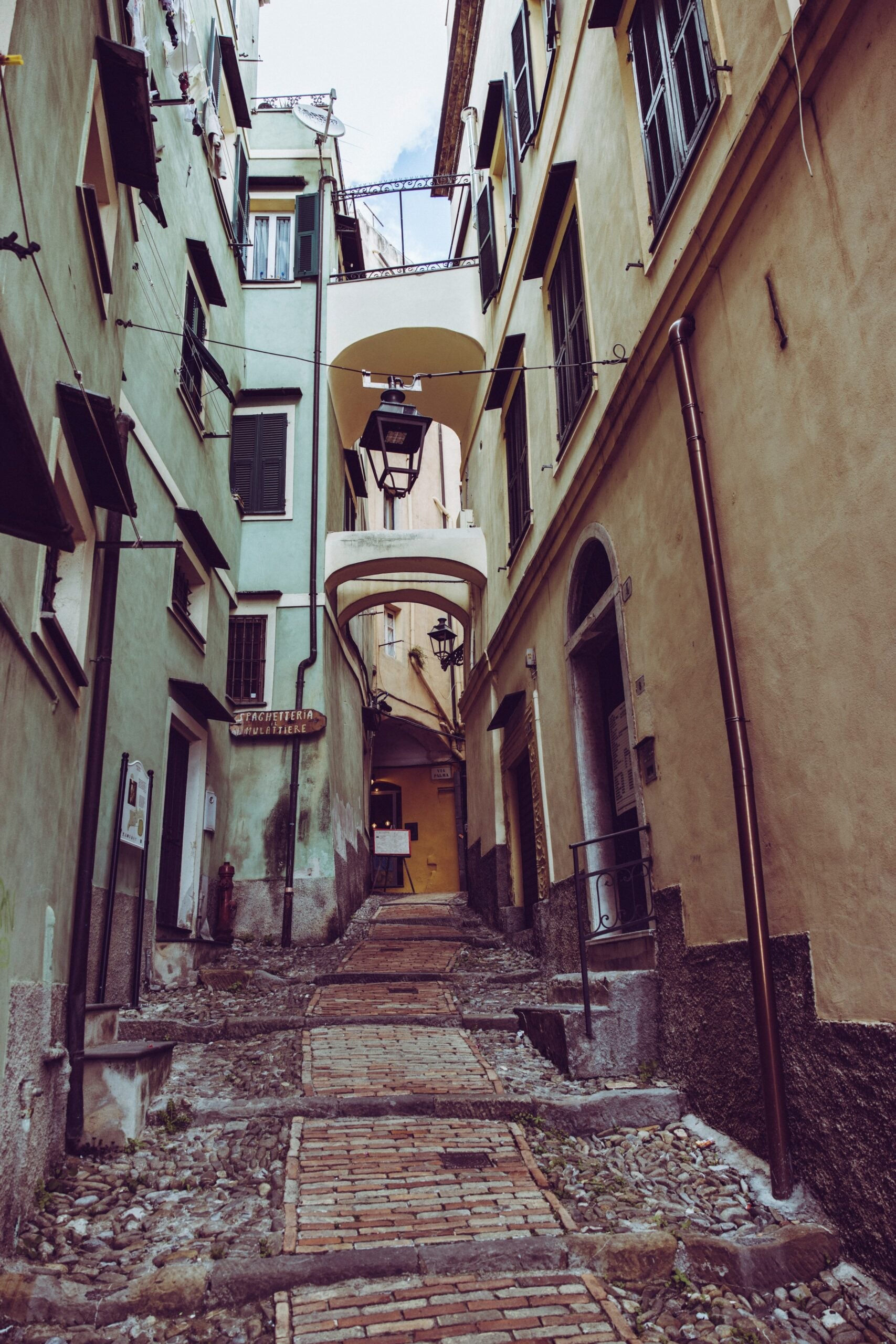 A quiet, narrow residential street in Sanremo, Italy, lined with old buildings that exude historic charm. The street, paved with worn cobblestones, winds gently between the closely spaced buildings, which feature colorful but slightly faded facades in shades of pastel yellow, pink, and beige. Shuttered windows, ornate balconies with wrought-iron railings, and hanging flower pots add to the quaint atmosphere. The street is empty, with no people or cars in sight, creating a serene, almost timeless ambiance. The scene captures the essence of everyday life in Sanremo, Italy, offering a glimpse into the town's tranquil and picturesque character.