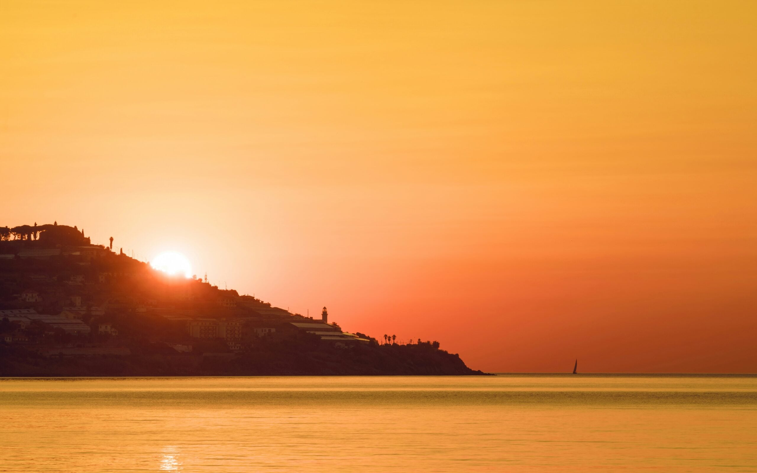 A breathtaking view of an orange-red sunset over the water in Sanremo, Italy. The sky is ablaze with vibrant hues of orange, red, and pink, casting a warm glow over the tranquil sea. The sun, partially dipping below the horizon, reflects beautifully on the calm waters, creating a shimmering pathway of light. Silhouettes of distant boats and the coastline add depth to the scene, highlighting the serene and romantic ambiance that Sanremo, Italy, is known for. This stunning sunset encapsulates the natural beauty and peaceful atmosphere of this Mediterranean coastal town.