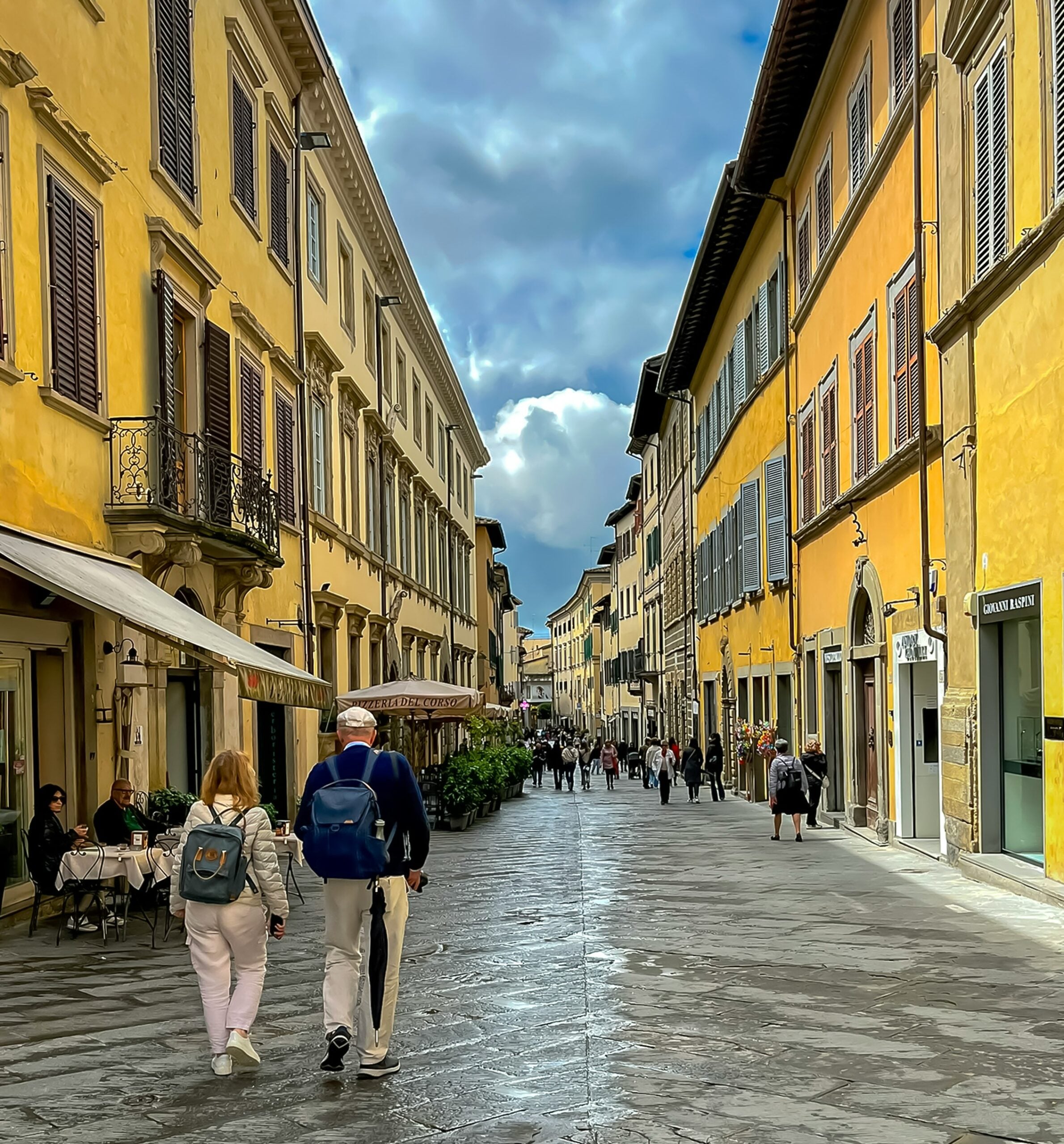 This image depicts a pedestrian street in downtown Arezzo, Italy, featuring a vibrant scene with people walking along the cobblestone path. The street is lined with charming yellow buildings that exude the warmth of Tuscan architecture, adding to the inviting ambiance of the area. The photograph captures the essence of daily life in Arezzo, Italy, showcasing the historic charm and lively atmosphere of this picturesque town, making it an ideal visual for travel guides or anyone interested in exploring the rich culture and scenic beauty of this Italian destination.