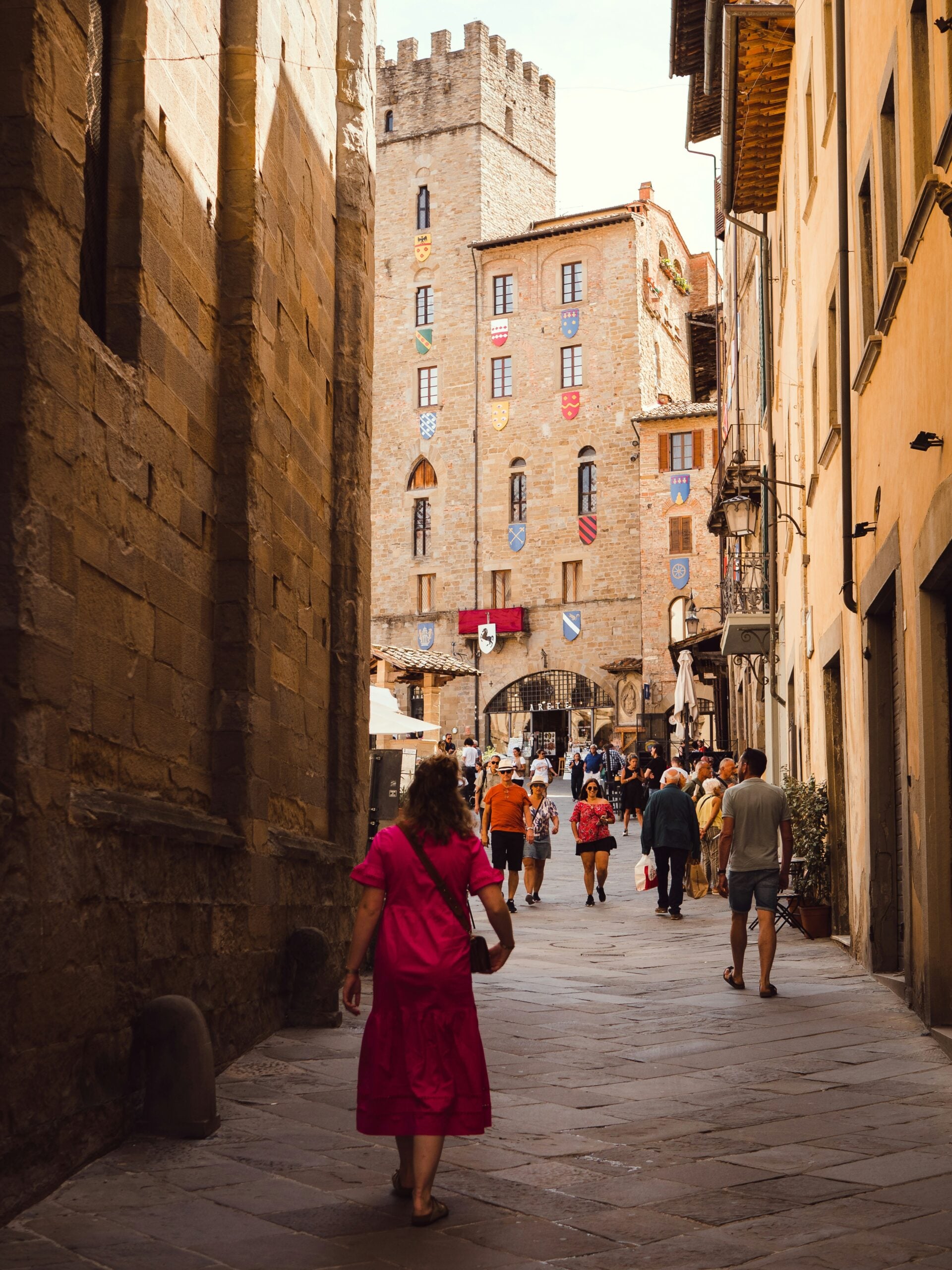The image depicts pedestrians walking along a narrow, cobblestone street in Arezzo, Italy, heading towards the town's main medieval piazza, Piazza Grande. The street is flanked by historic buildings with rustic facades, wooden shutters, and flower-filled balconies, creating a charming, old-world ambiance. The pathway gently slopes upward, leading toward the open space of the piazza, which is hinted at in the distance. The scene is lively yet intimate, with the pedestrians adding a sense of movement and daily life to the picturesque setting. This view captures the essence of Arezzo's medieval charm, highlighting the town's rich history and inviting atmosphere.