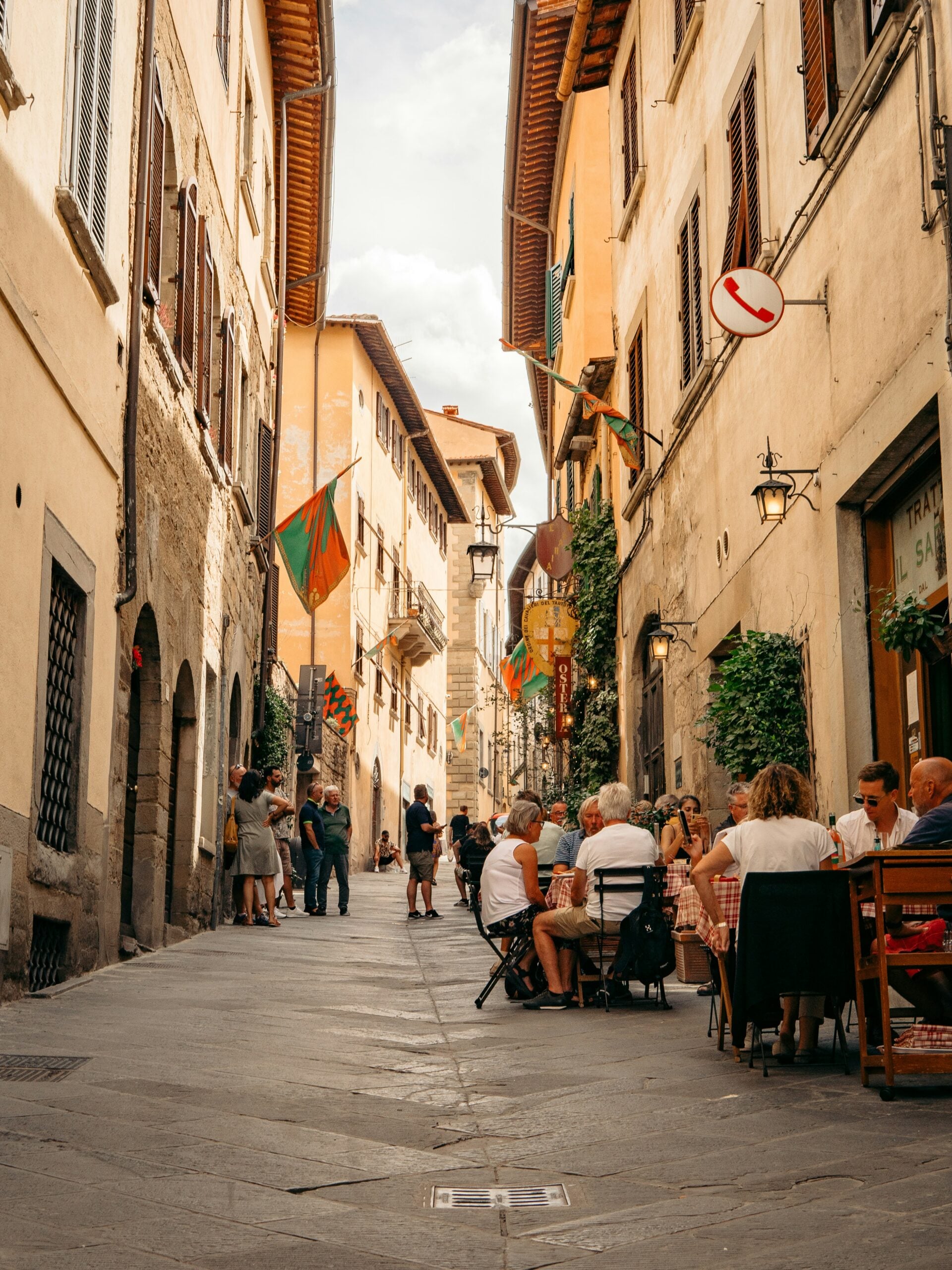 The image depicts a lively scene of people enjoying lunch outside on a pedestrian street in downtown Arezzo, Italy. Diners are seated at quaint outdoor tables beneath colorful umbrellas, surrounded by the charming architecture of historic buildings. The atmosphere is vibrant yet relaxed, with the cobblestone street adding to the picturesque setting. Passersby stroll by, contributing to the dynamic street life typical of Arezzo. The warm, inviting ambiance of this downtown area highlights the town's rich cultural atmosphere, making it an ideal spot for savoring Italian cuisine while soaking in the beauty of Arezzo, Italy.