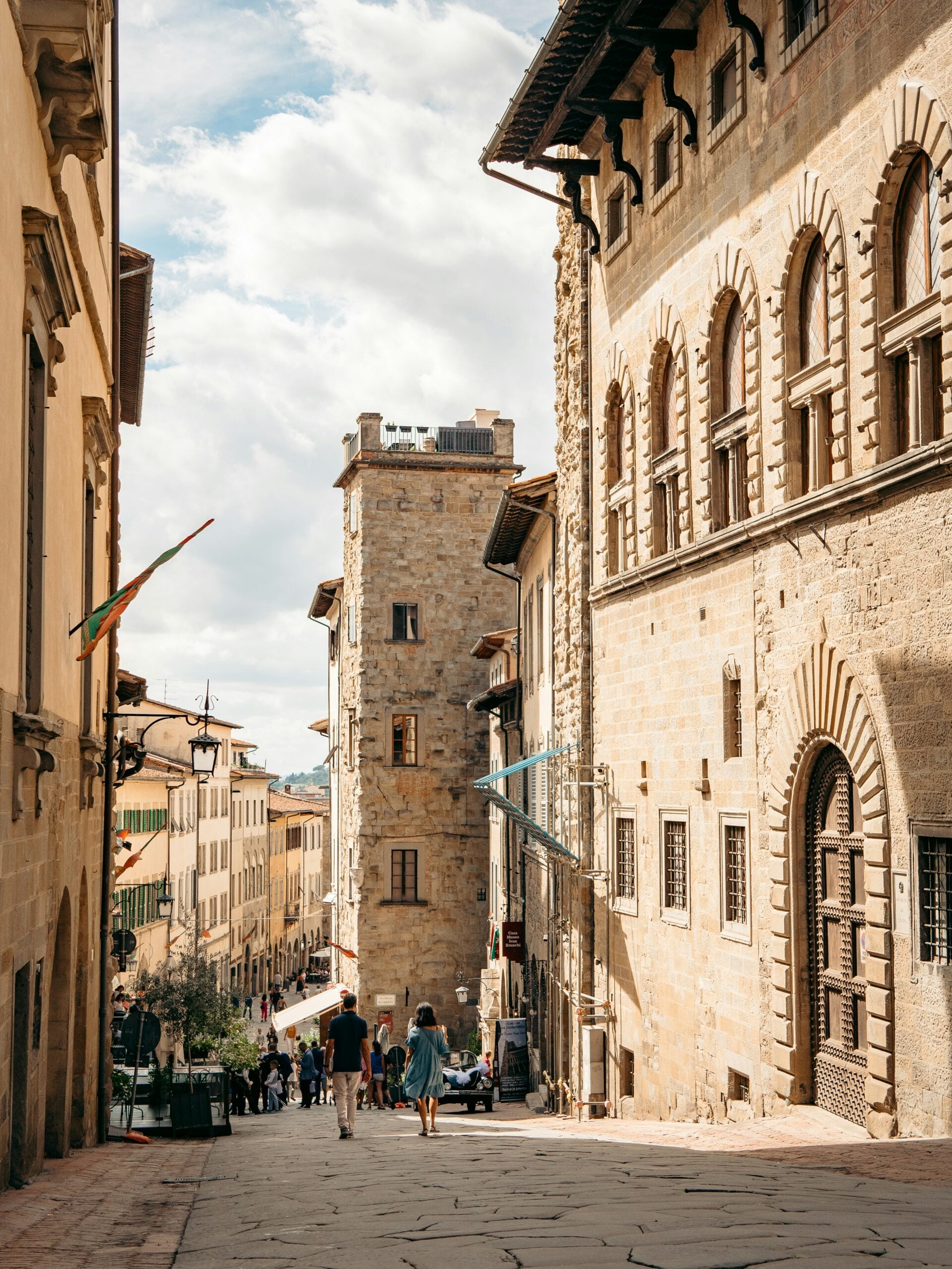 The image depicts people walking downhill on a pedestrian street in the old medieval section of Arezzo, Italy. The narrow cobblestone street is lined with historic buildings featuring stone facades, arched doorways, and wooden shutters, all reflecting the town's rich medieval heritage. The street slopes gently, leading toward a lower part of the town, with the walkers adding a sense of movement and life to the scene. The atmosphere is both lively and serene, with the timeless architecture providing a picturesque backdrop. This image beautifully captures the essence of daily life in the medieval heart of Arezzo, Italy.