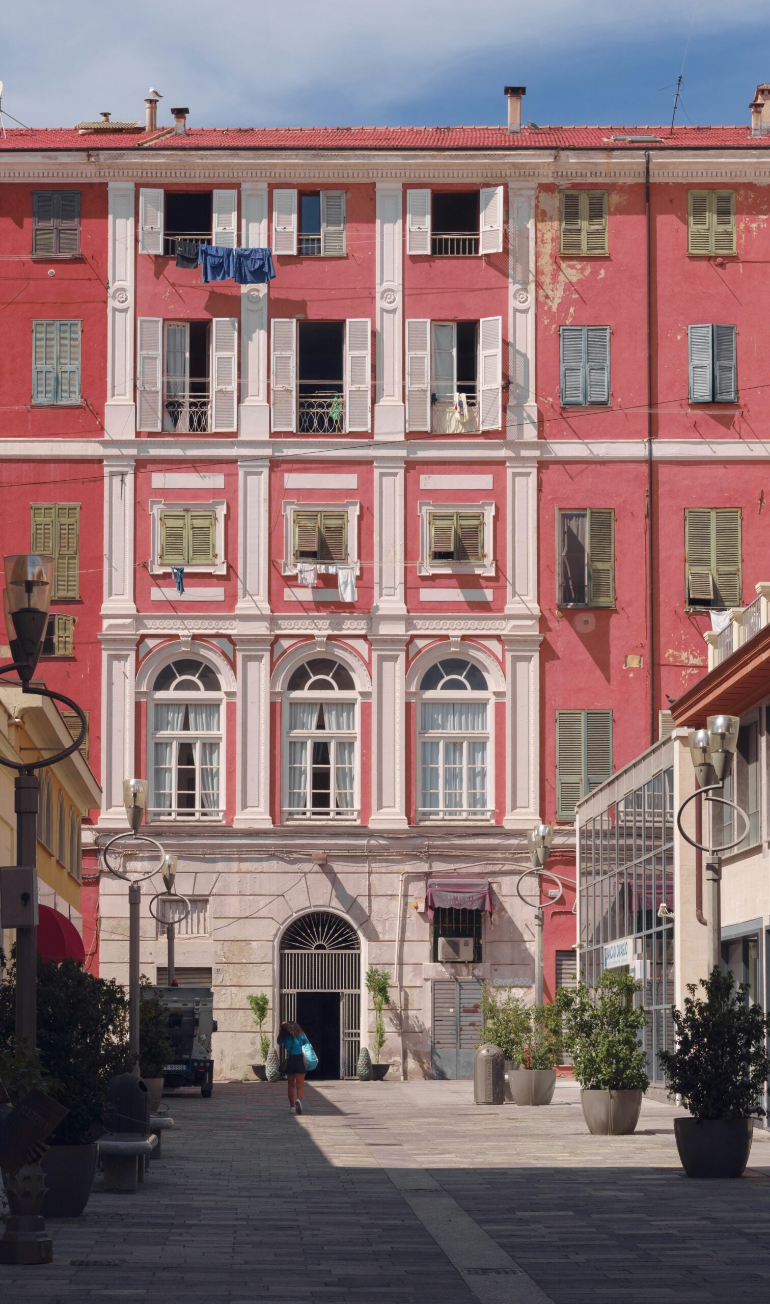 A charming pink residential building in Sanremo, Italy, showcasing the region's classic Mediterranean architecture. The building features ornate balconies with wrought-iron railings, vibrant green shutters, and decorative details that add character to its pastel facade. Set against a backdrop of clear blue skies, the pink hue of the building stands out, reflecting the colorful and lively spirit of Sanremo, Italy. This picturesque scene captures the essence of everyday life in this coastal town, where vibrant architecture and scenic surroundings create a uniquely inviting atmosphere.