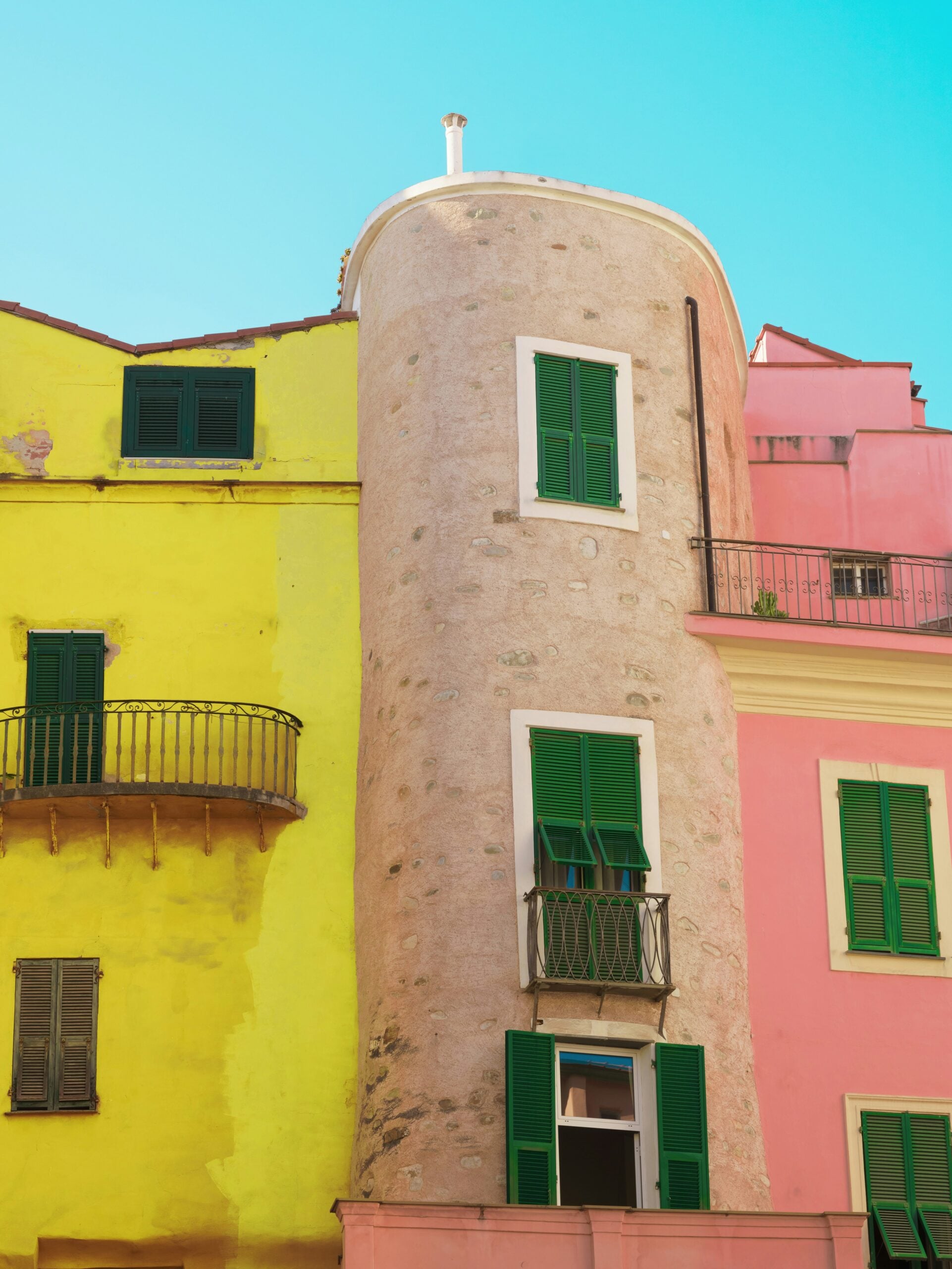 A vibrant residential building facade in Sanremo, Italy, painted in alternating hues of yellow and pink. The building's exterior exudes a lively and cheerful atmosphere, with the contrasting colors creating a visually striking effect. Each window is adorned with classic green shutters, which add a touch of traditional Mediterranean charm to the overall aesthetic. The building's architectural details, including ornate balconies and decorative trim, further enhance its character. Set against a backdrop of clear blue skies, this colorful facade captures the essence of Sanremo's coastal charm and vibrant lifestyle.