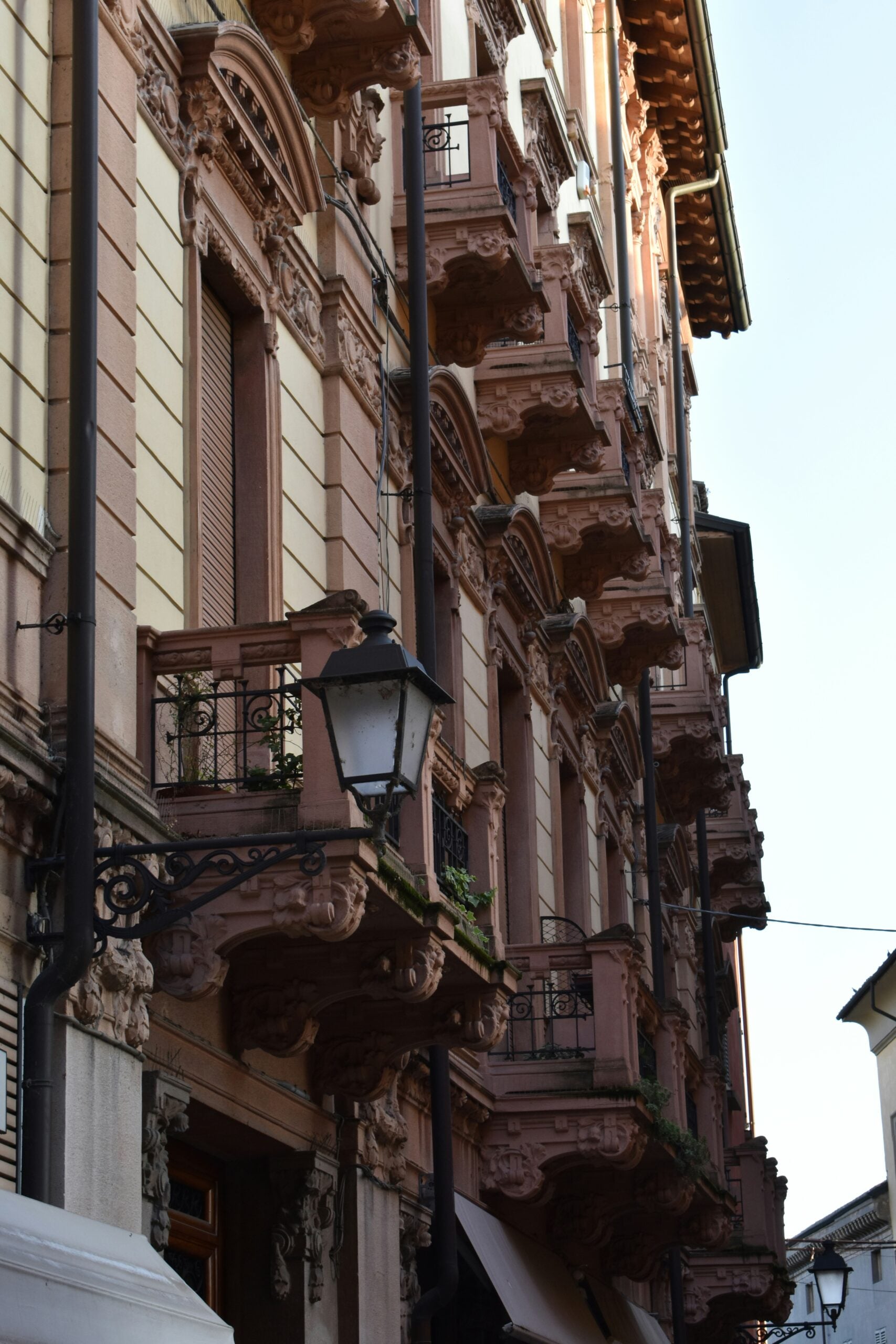 A photograph of the facade of residential buildings in Reggio Emilia, Province of Reggio Emilia, Italy, showcases the region's characteristic architectural style. The buildings feature warm, earthy tones, with pastel-colored walls that range from soft yellows to muted terracottas. Shuttered windows, some with flower boxes brimming with vibrant blooms, add to the charm of these homes. The facades are adorned with intricate wrought-iron balconies and arched doorways, reflecting the classic Italian design that defines this historic area. This image beautifully captures the quaint, yet elegant, essence of residential life in Reggio Emilia, Italy.