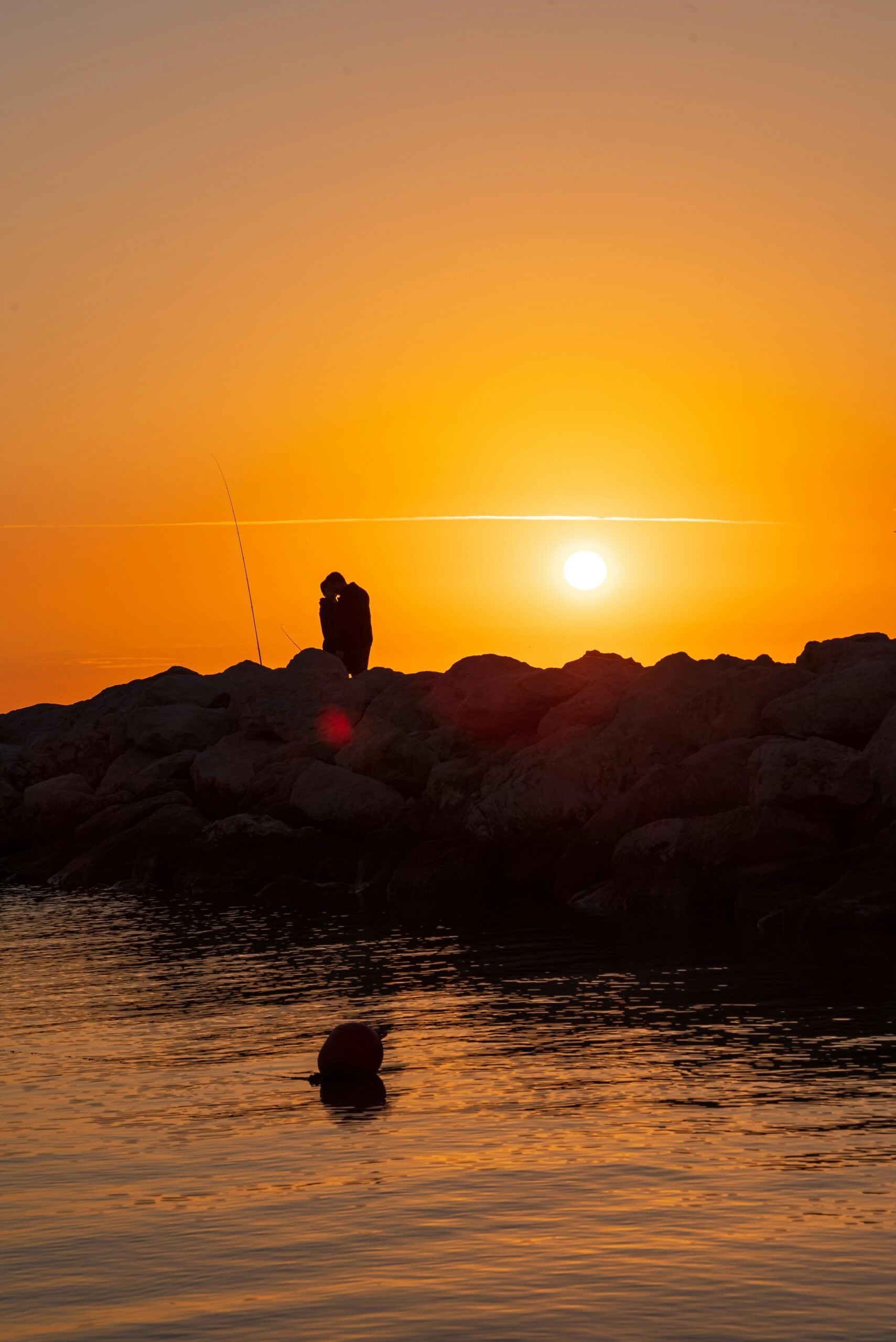 A romantic silhouette of a couple kissing at sunset on a dock in Pescara, Italy. The couple stands at the edge of the dock, their outlines dark against the backdrop of a vibrant sky painted with warm oranges, pinks, and purples as the sun dips below the horizon. The calm Adriatic Sea reflects the stunning colors, creating a mirror-like surface that enhances the serene atmosphere. The iconic skyline of Pescara, including distant mountains and coastal elements, subtly frames the scene, adding a sense of place to this intimate and timeless moment.