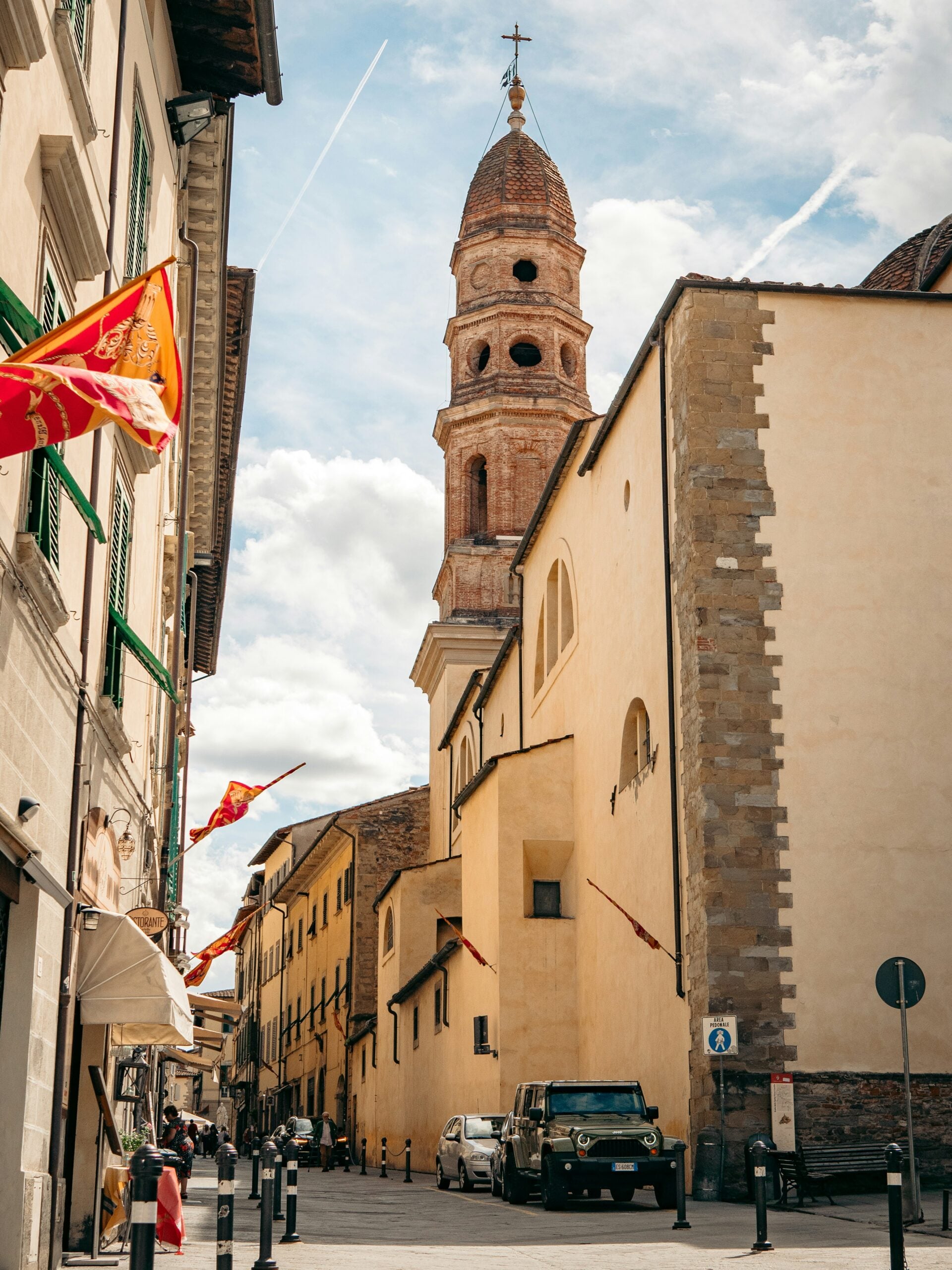 The image showcases a charming street in Arezzo, Italy, with a picturesque view of a church tower rising in the background. The street is lined with historic buildings featuring warm, earthy tones typical of Tuscan architecture, with arched doorways and shuttered windows. The cobblestone pathway leads the eye toward the prominent church tower, which stands tall against a clear blue sky, adding a sense of history and grandeur to the scene. The tranquil atmosphere of the street, combined with the striking presence of the church tower, captures the timeless beauty and rich cultural heritage of Arezzo, Italy.