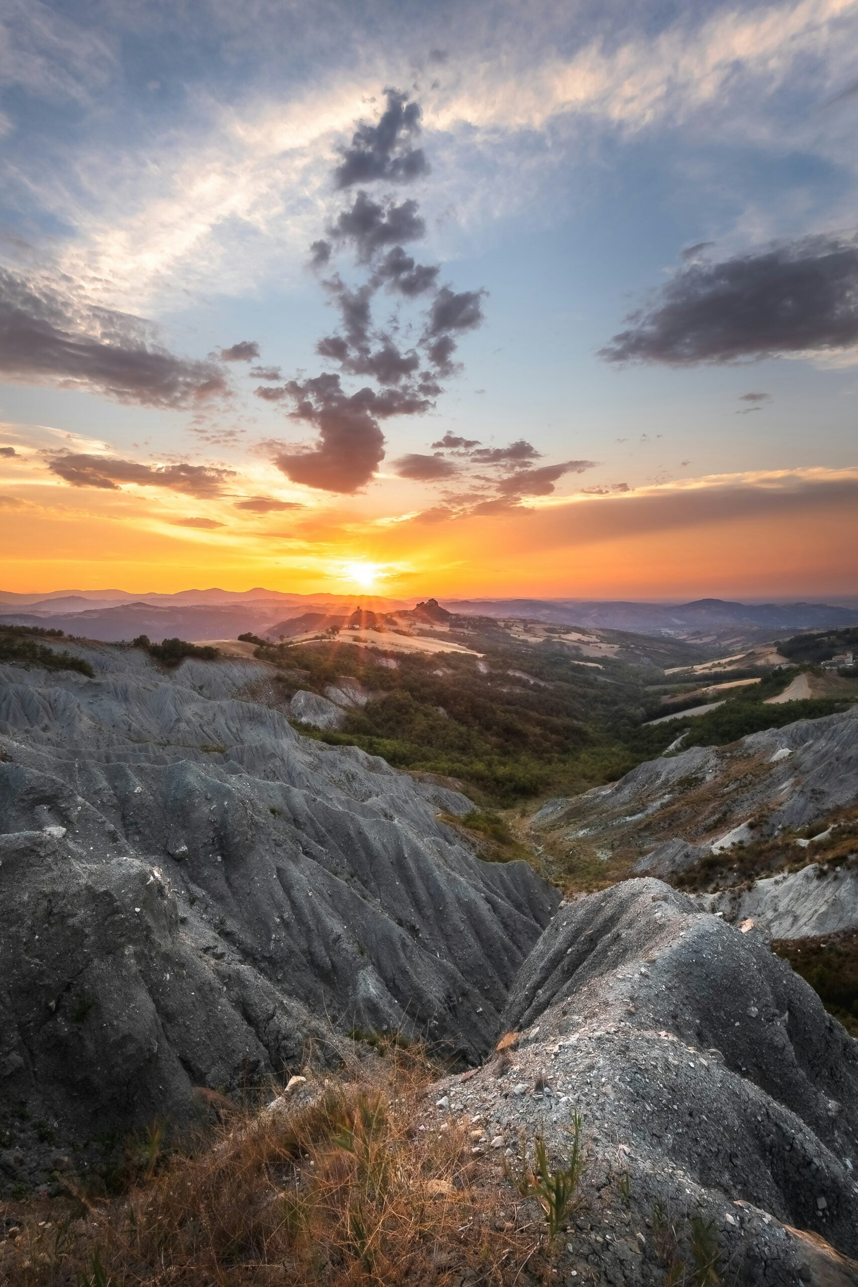 A photograph captures the serene beauty of a sunrise landscape in Rossena, Province of Reggio Emilia, Italy. The early morning light bathes the rolling hills in a soft, golden glow, highlighting the lush greenery and the silhouette of a distant medieval castle perched on a hilltop. The sky is a canvas of warm hues, transitioning from deep oranges to soft pinks and purples, reflecting off the gentle mist that blankets the valley. This tranquil scene encapsulates the timeless beauty of the Italian countryside, inviting viewers to experience the peacefulness and natural splendor of Rossena, Reggio Emilia.
