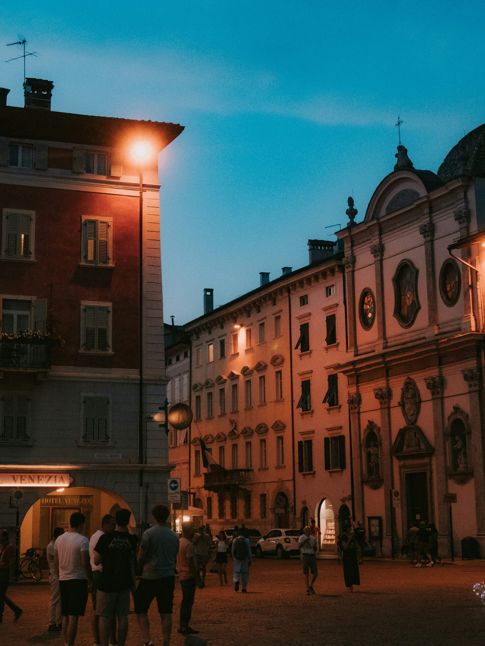 A picturesque evening scene of a bustling piazza in Trento, Italy, during a warm summer night. The square is alive with the soft glow of streetlights and the ambient light from surrounding cafes and restaurants. People are enjoying leisurely strolls, dining al fresco, and socializing, while the historic architecture of the piazza, with its ornate facades and charming details, adds to the enchanting atmosphere. The sky is tinged with the last hues of sunset, casting a warm, golden light over the scene. This vibrant evening captures the lively yet relaxed spirit of summer in Trento, Italy.