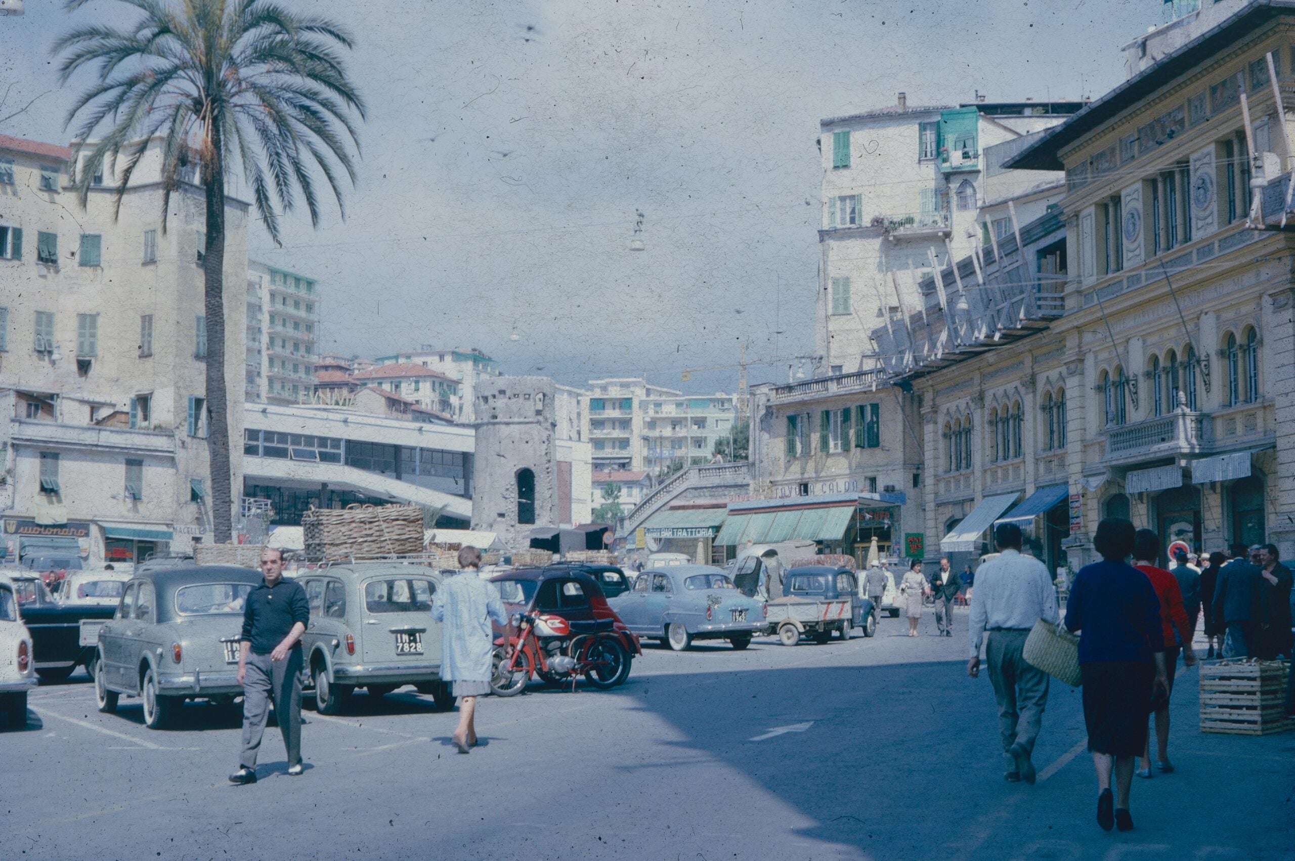 A nostalgic, color-faded photograph of a vintage piazza scene in Sanremo, Italy. The image captures the essence of a bygone era, with people leisurely strolling across the cobblestone square, surrounded by charming, weathered buildings. The architecture, typical of the region, features arched doorways, pastel-colored facades, and rustic balconies. In the center of the piazza, a fountain or statue serves as a focal point, where locals and visitors gather. The colors have softened over time, giving the photo a dreamy, timeless quality that evokes the serene and historic ambiance of Sanremo, Italy, in its earlier days.