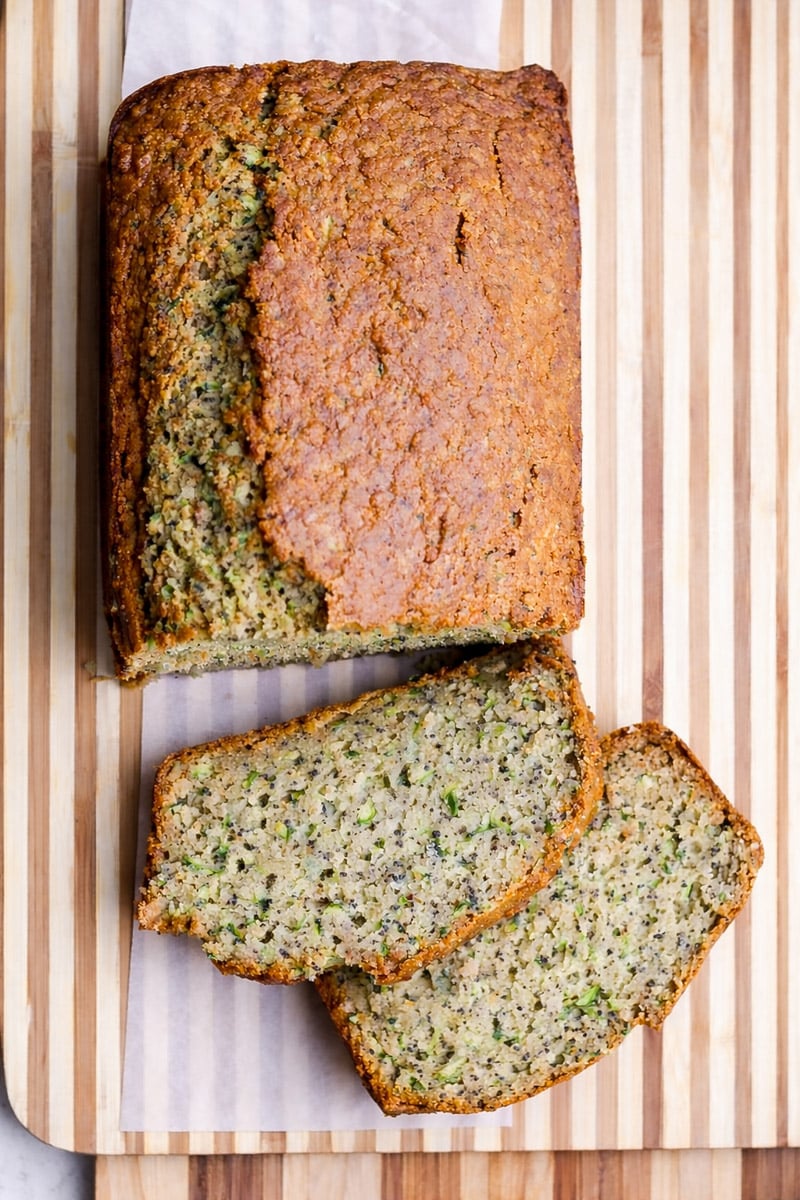 A loaf of healthy zucchini bread sits on a striped wooden cutting board, with two slices cut to reveal a moist, speckled interior and visible green zucchini pieces.