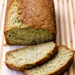 A loaf of healthy zucchini bread sits on a striped wooden cutting board, with two slices cut and lying in front, showing a moist, speckled interior with visible green zucchini flecks.