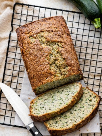 A loaf of healthy zucchini bread sits on a cooling rack, partially sliced with two pieces cut. A knife rests beside the bread, and a whole zucchini is visible in the top right corner.