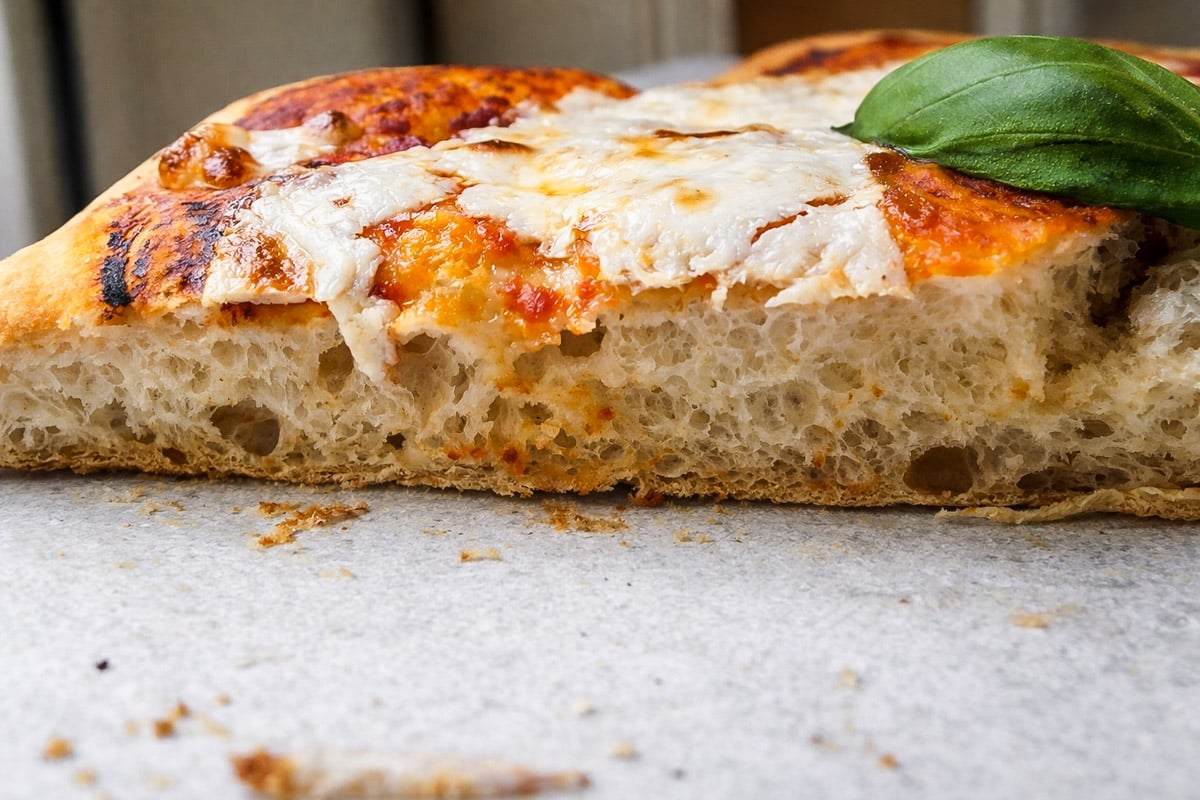 A close-up of a Sourdough Pizza Margherita slice shows a thick, airy crust with melted cheese, tomato sauce, and a green basil leaf on top.