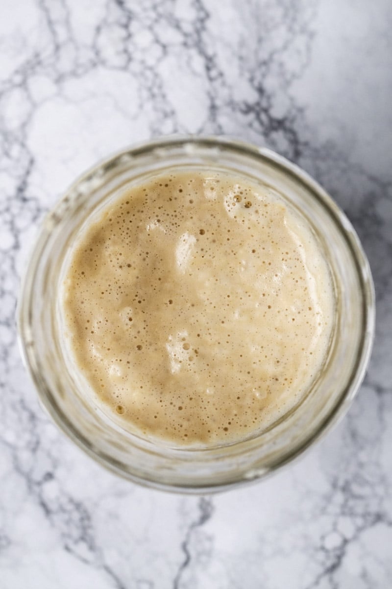 A glass jar filled with bubbly, active sourdough starter&mdash;perfect for Sourdough Pizza Margherita&mdash;sits on a white marble surface, viewed from above. The starter has a creamy tan color and a foamy texture with visible air bubbles.