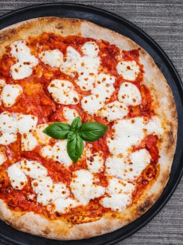 A Sourdough Pizza Margherita topped with melted mozzarella cheese, tomato sauce, and fresh basil leaves, served on a round black tray atop a textured gray surface.