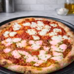 A freshly baked Sourdough Pizza Margherita with a thick, bubbly crust, topped with melted mozzarella cheese and tomato sauce, sits on a round baking tray on a marble countertop. Kitchen utensils and olive oil are in the background.