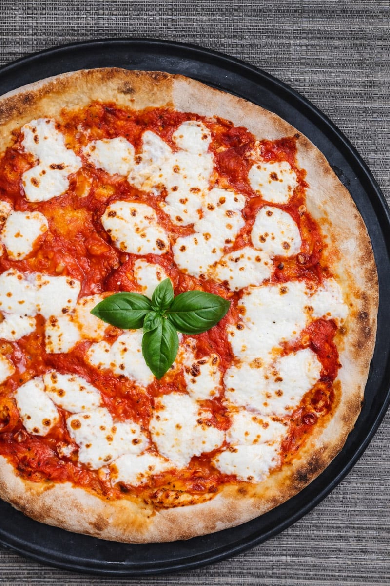 A Sourdough Pizza Margherita topped with melted mozzarella cheese, tomato sauce, and fresh basil leaves, served on a round black tray atop a textured gray surface.