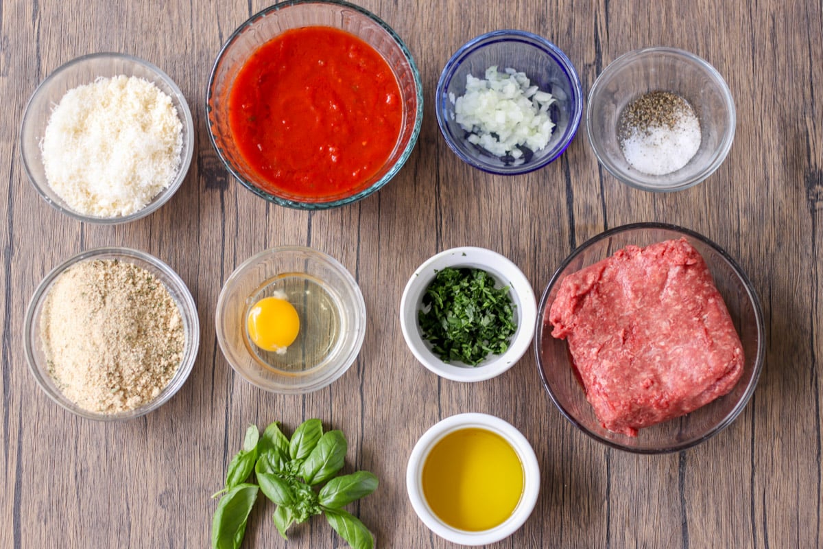 overhead ingredients to make meatballs in glass bowls plus tomato sauce and basil.