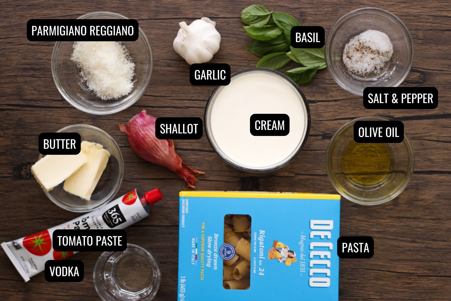 Ingredients for a classic rigatoni alla vodka pasta dish, including pasta, olive oil, pepper, basil, garlic, cream, shallot, butter, tomato paste, vodka, and grated Parmigiano Reggiano arranged on a wooden surface.