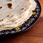 A stack of flour tortillas, reminiscent of piadina Italian flatbread, sits on a dark, decorative plate with floral patterns, placed on a wooden table.