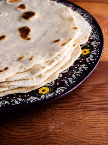 A stack of flour tortillas, reminiscent of piadina Italian flatbread, sits on a dark, decorative plate with floral patterns, placed on a wooden table.