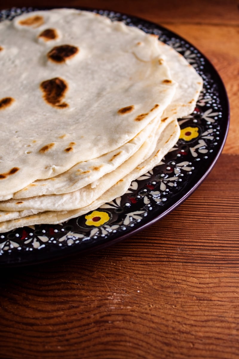 A stack of flour tortillas, reminiscent of piadina Italian flatbread, sits on a dark, decorative plate with floral patterns, placed on a wooden table.