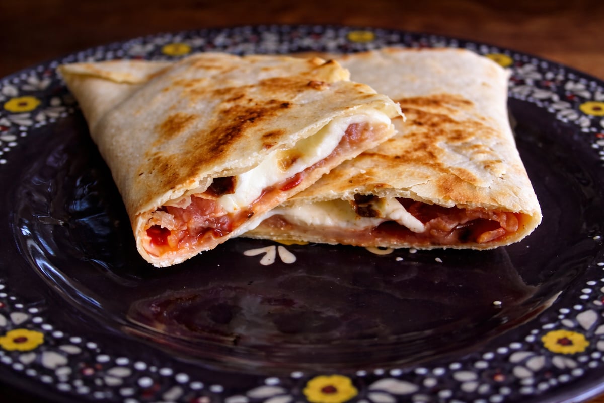 A close-up of a sliced calzone filled with melted cheese and tomato sauce, served on a decorative dark plate with floral patterns, reminiscent of piadina Italian flatbread tradition.