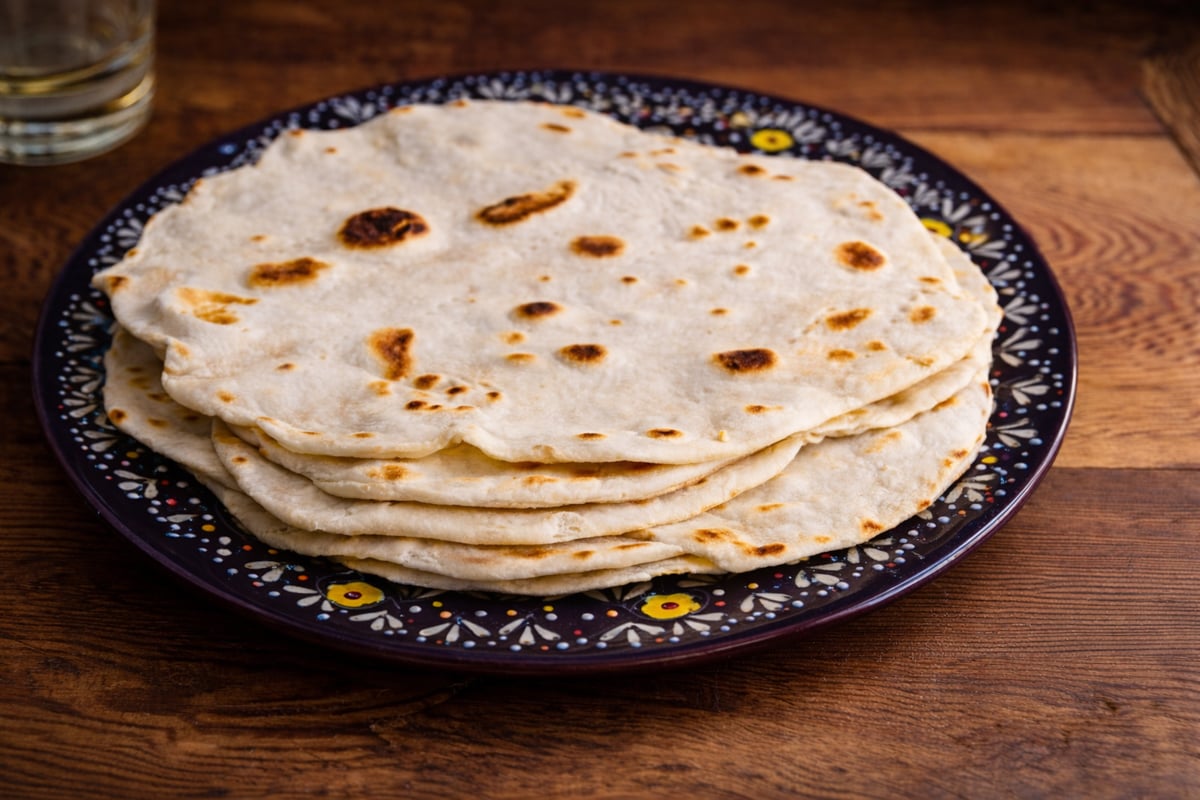 A stack of homemade flour tortillas, reminiscent of piadina Italian flatbread, sits on a decorative dark plate with white and yellow patterns, placed on a wooden table.