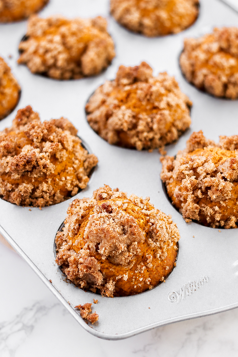 A metal muffin tin containing pumpkin muffins topped with brown sugar crumble.