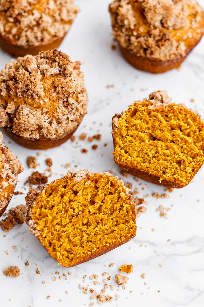 Several pumpkin muffins with crumb toppings, including one cut in half to show the golden orange interior.