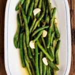 An oval white platter filled with sautéed green beans and garlic cloves on a wooden surface.