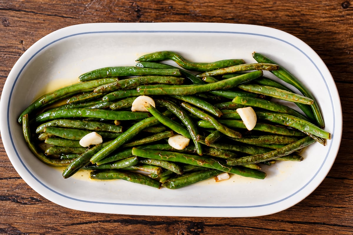 Oval platter of sautéed green beans with garlic cloves on a wooden table.