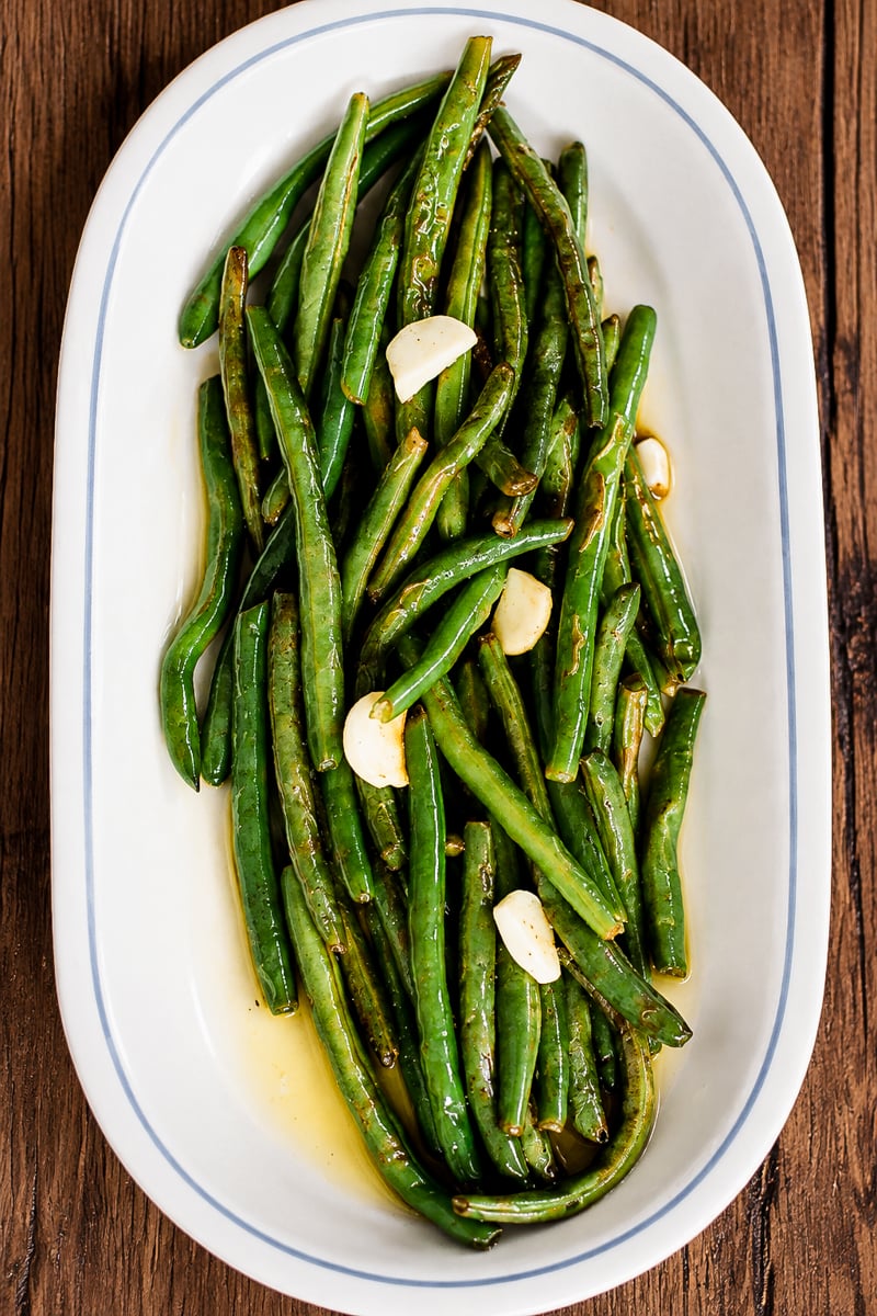An oval white platter filled with sautéed green beans and garlic cloves on a wooden surface.