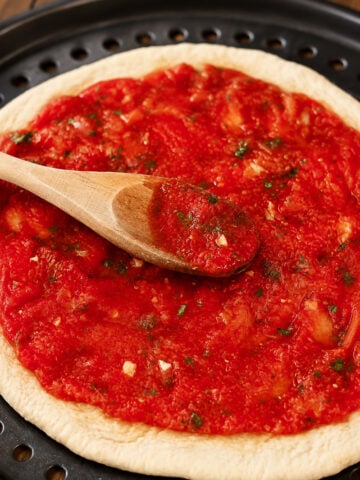 A wooden spoon swirls rich tomato sauce over a round, uncooked pizza crust on a perforated pizza pan.