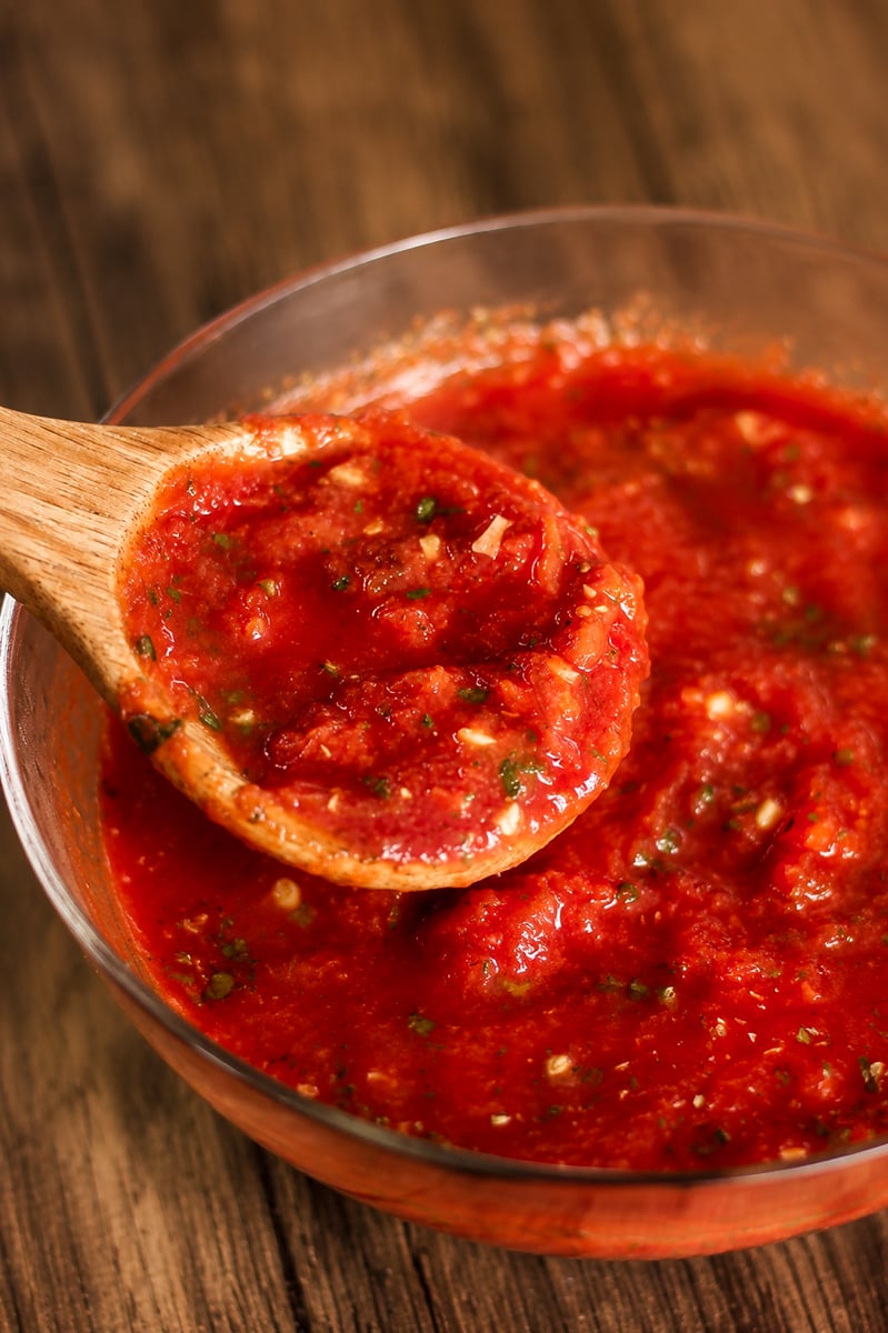 A wooden spoon holds up a serving of chunky tomato sauce, rich with herbs, from a clear glass bowl on a wooden surface.