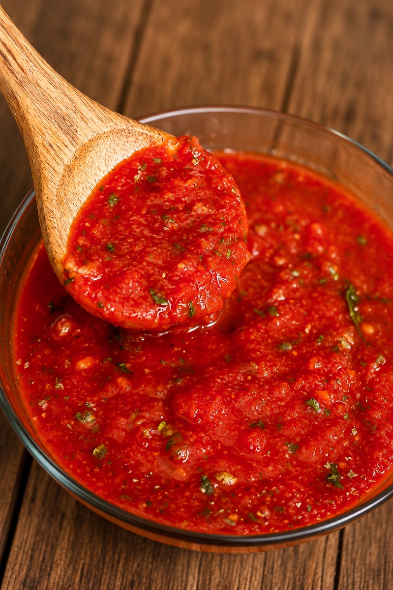 A wooden spoon holding chunky red tomato salsa, reminiscent of fresh tomato sauce, hovers above a glass bowl on a wooden table. The salsa contains visible bits of herbs and tomatoes.