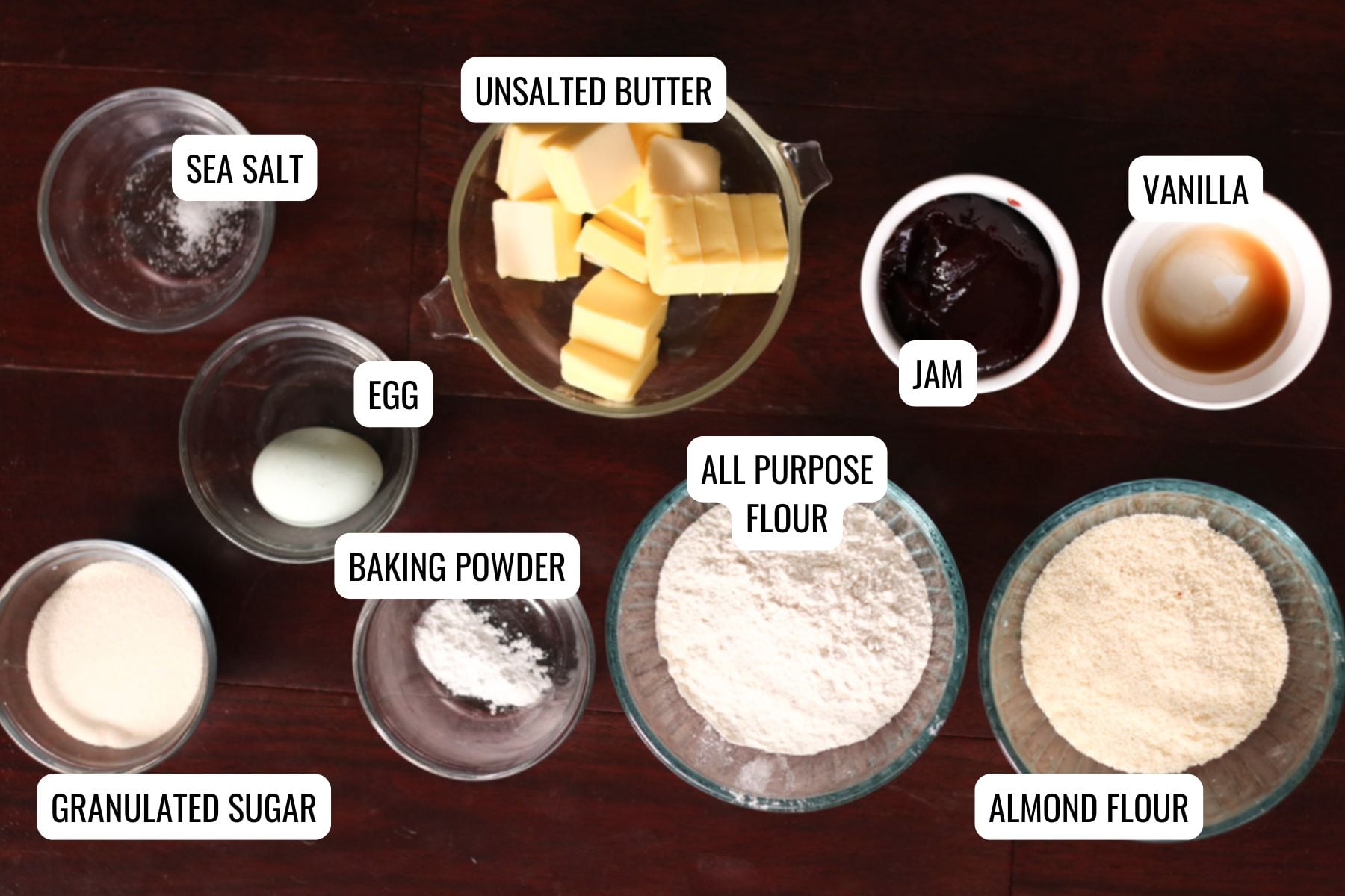 A flat lay of baking ingredients in bowls on a dark surface, labeled: sea salt, an egg, granulated sugar, baking powder, unsalted butter, all purpose flour, almond flour, jam, and vanilla—perfect for making thumbprint cookies.
