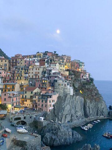 Colorful cliffside buildings overlook a small harbor with boats in Cinque Terre, set against a rocky coastline and calm blue sea at dusk. The sky is pale with a bright moon shining above the picturesque village.