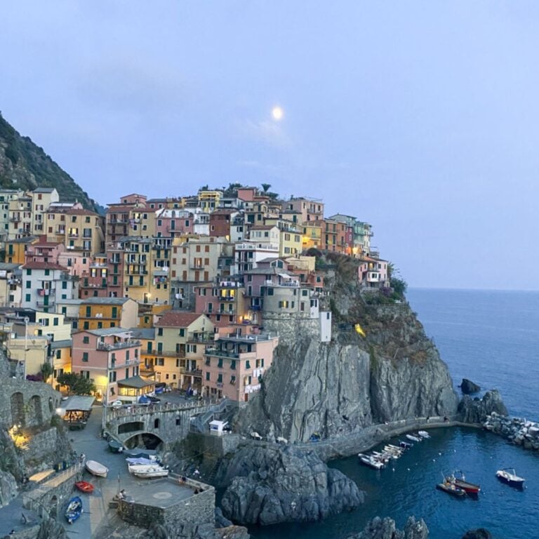Colorful cliffside buildings overlook a small harbor with boats in Cinque Terre, set against a rocky coastline and calm blue sea at dusk. The sky is pale with a bright moon shining above the picturesque village.
