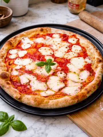 An easy homemade pizza, this freshly baked margherita features melted mozzarella, tomato sauce, and basil atop a black tray on a marble countertop. A sprig of basil centers the pie among kitchen tools and ingredients.
