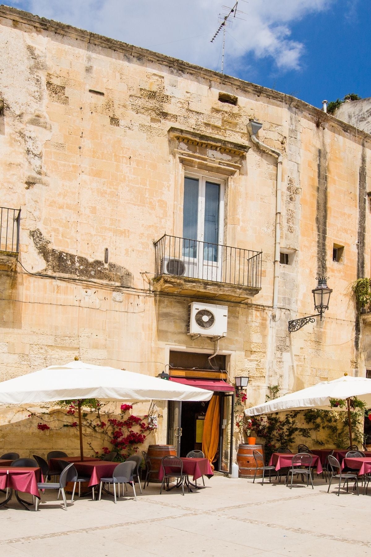 A rustic stone building in Lecce with a small balcony, potted plants, and an air conditioner. Outdoor tables with red tablecloths and white umbrellas invite you to this cozy cafe or restaurant.