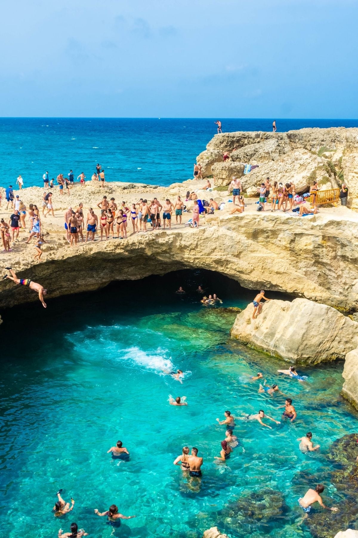 Tourists swim and relax in and around a natural rock pool with clear turquoise water by the sea near Lecce; some people jump from the cliffs while others watch or swim below, under a bright blue sky.
