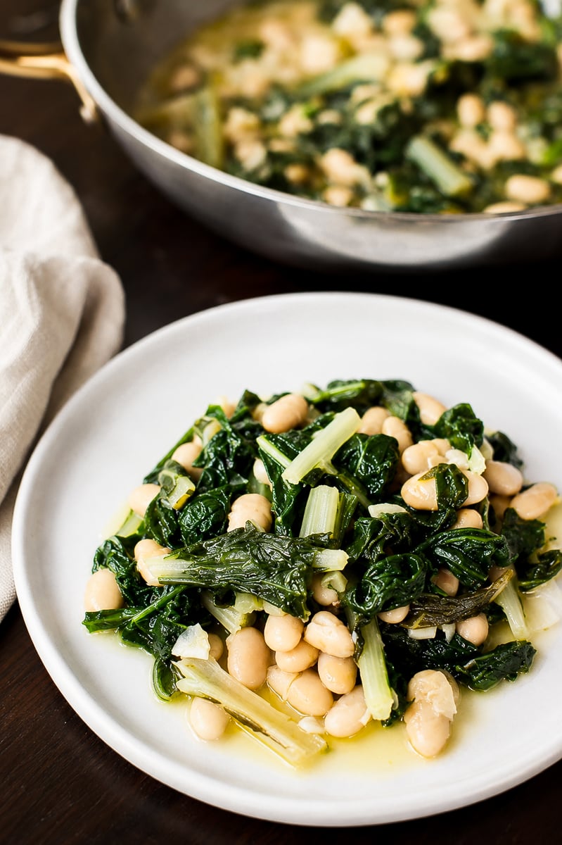 A white plate with sautéed greens—featuring rainbow chard—and white beans in olive oil, with a pan containing more of the dish in the background.