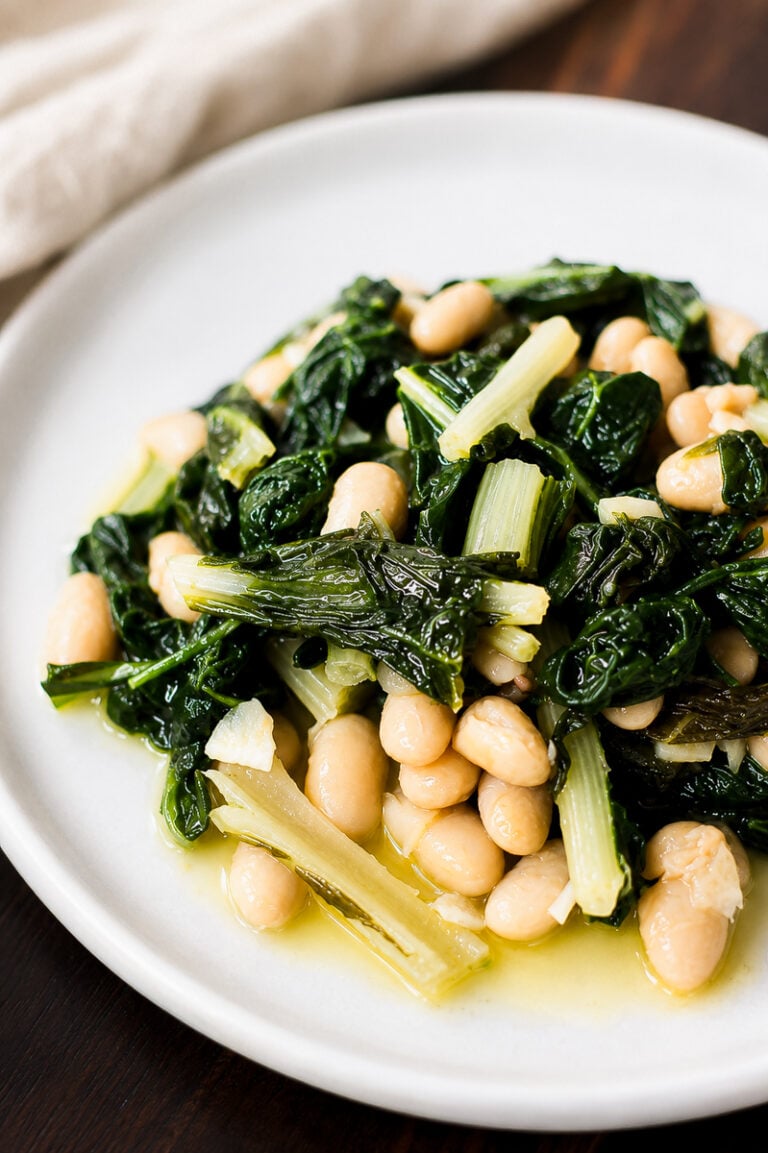 A white plate with sautéed rainbow chard and white beans in olive oil, served on a dark wooden table.
